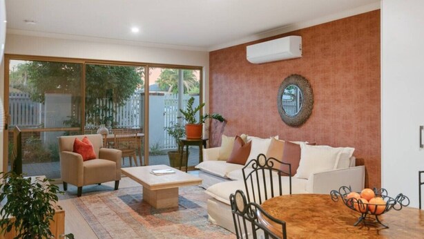 A living room with a sofa, armchair, coffee table, and a dining table with chairs. There is a large window with a view of a fence and trees. - Terra de Sol | Terracotta Courtyard, Swan Valley (Bassendean)