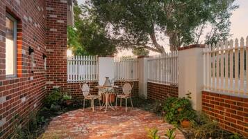 A brick patio with a table and chairs, surrounded by a white picket fence and greenery.