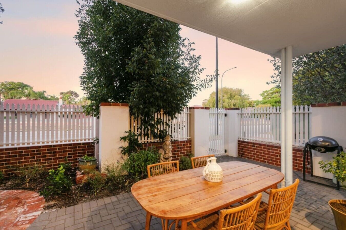 An outdoor patio with a wooden table and chairs, surrounded by a white fence and greenery.