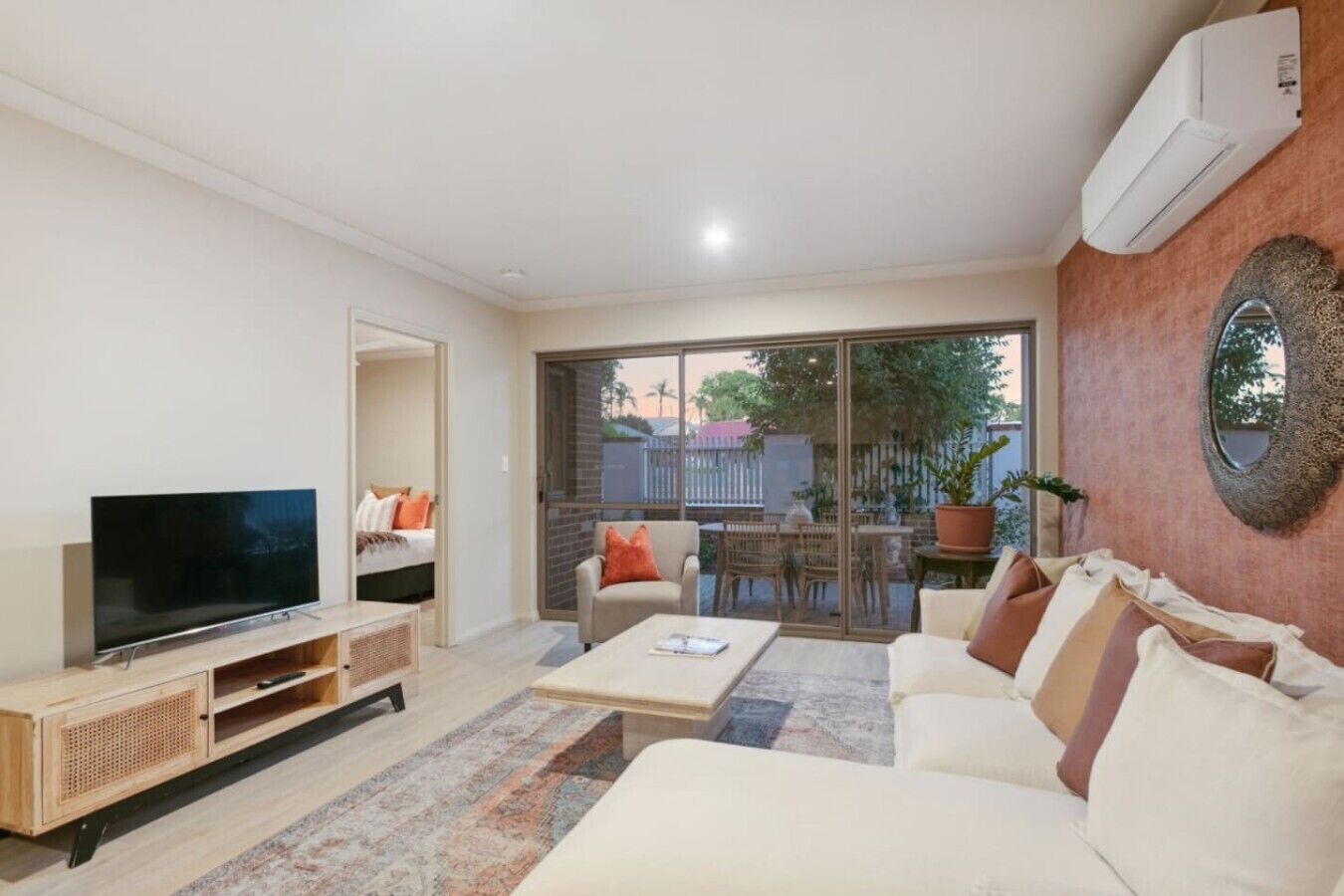 A modern living room with a flat-screen TV, a sofa, a coffee table, and a dining area visible through glass doors.