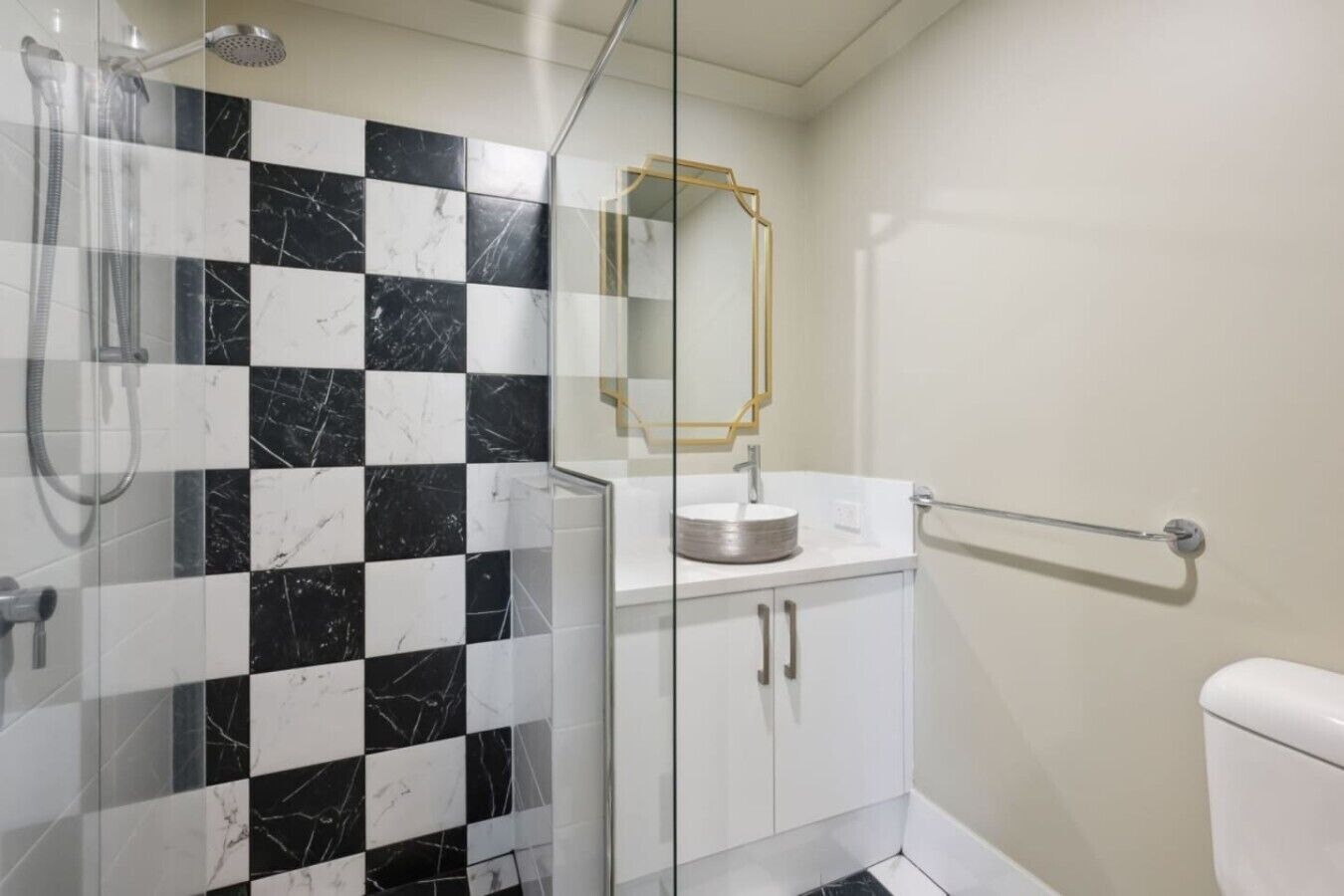 A bathroom with a checkered black and white tile wall, a glass shower enclosure, a white sink with a gold-framed mirror, and a white toilet.