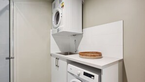 A compact laundry room with a front-loading washer and dryer, a small sink, and a basket on the countertop. - The Tonic | Teal, Timber & Courtyard, Swan Valley (Bassendean)