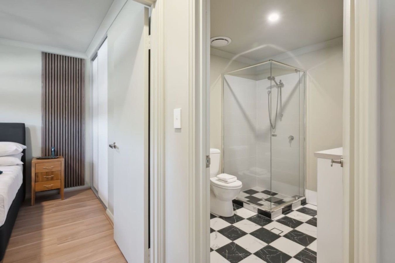 A hotel room with a bed, a wooden nightstand, a bathroom with a glass-enclosed shower, a toilet, and a checkered floor.