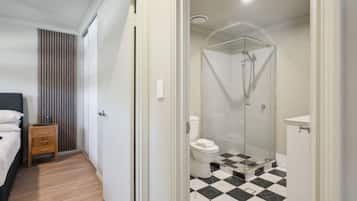 A hotel room with a bed, a wooden nightstand, a bathroom with a glass-enclosed shower, a toilet, and a checkered floor.