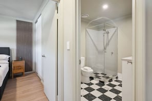 A hotel room with a bed, a wooden nightstand, a bathroom with a glass-enclosed shower, a toilet, and a checkered floor. - The Tonic | Teal, Timber & Courtyard, Swan Valley (Bassendean)