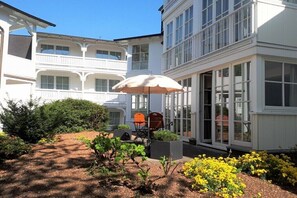 A two-story building with a balcony, a covered seating area with an umbrella, and a garden with yellow flowers. - Design Apartment Ambiente (Ostseebad Binz)