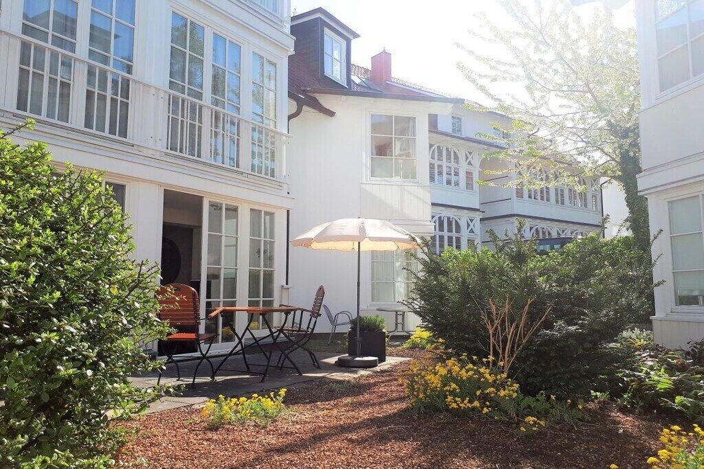 A courtyard with a table and chairs, surrounded by greenery and a white building with large windows.