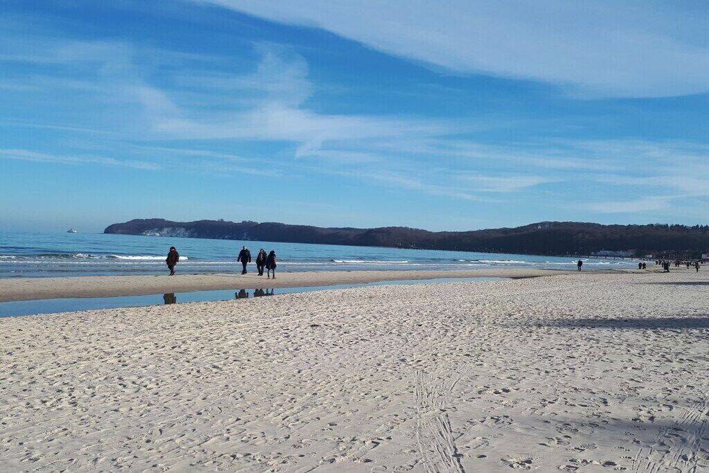 A beach with people walking, a boat in the water, and a hilly landscape in the distance.