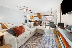 A modern living room with a sectional sofa, a patterned rug, a ceiling fan, and a kitchen area visible through an open door. - Bliss Retreat Whitefish Studio (Whitefish)