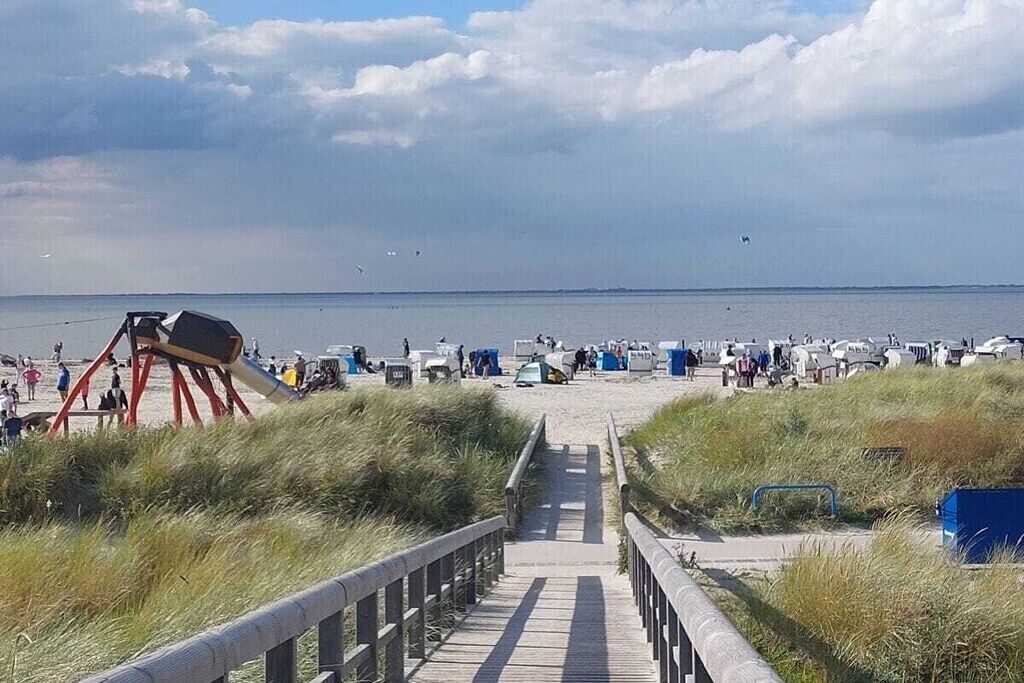 A beach with a wooden walkway leading to the water, surrounded by dunes and a clear sky.
