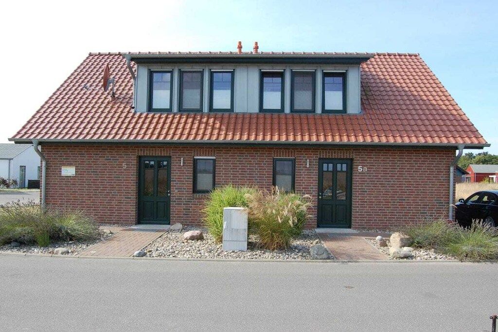 A two-story brick building with a red-tiled roof, two green doors, and a small garden area with rocks and plants.