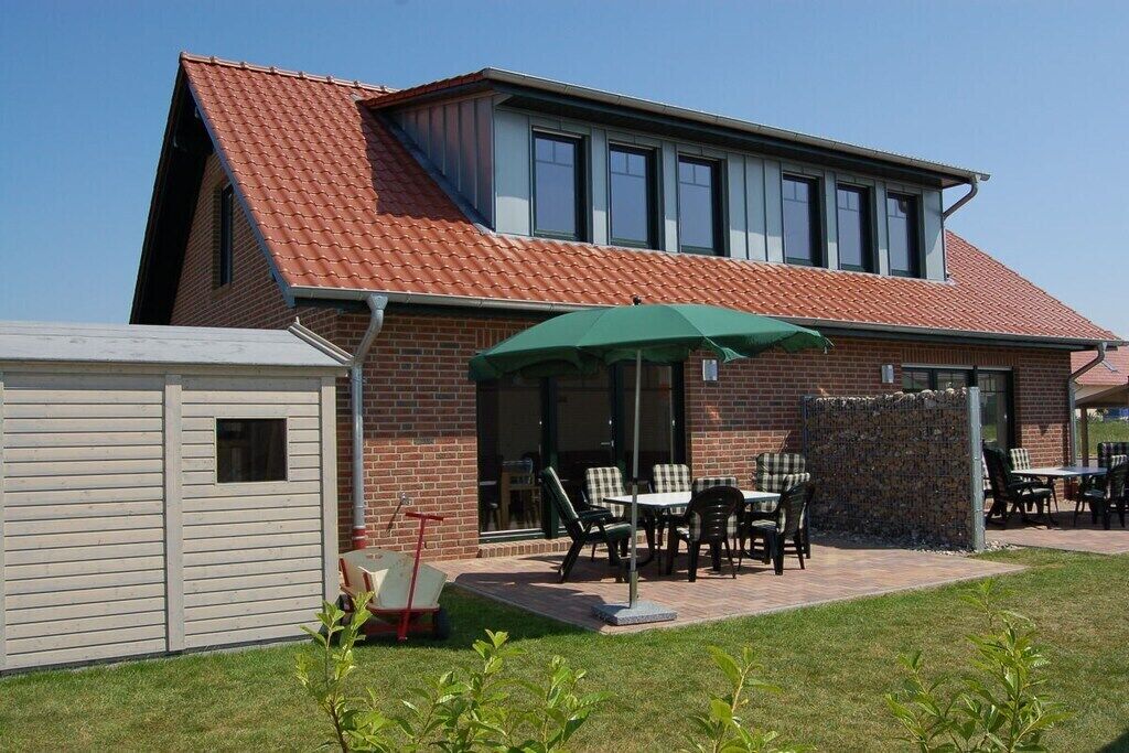 Une maison à deux étages avec un toit recouvert de tuiles rouges, une terrasse couverte avec des meubles d’extérieur et un petit abri de jardin.