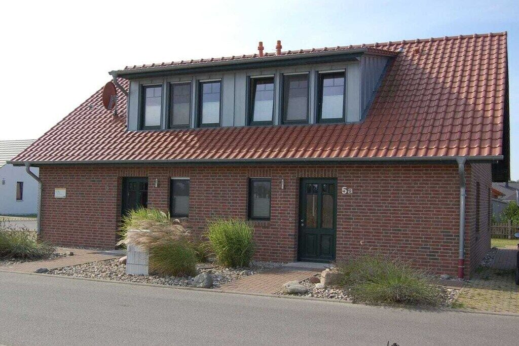 Ein zweistöckiges Backsteingebäude mit rotem Ziegeldach, einer grünen Tür und einem kleinen Gartenbereich mit Steinen und Pflanzen.