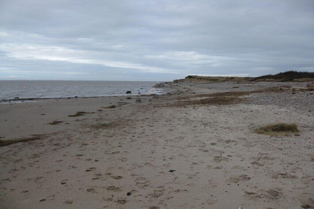 A beach with a wide sandy area, a rocky outcrop, and a distant view of the sea under a cloudy sky.