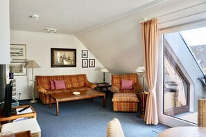 A cozy living room with a sofa, armchair, and coffee table. There is a window with curtains, a picture on the wall, and a lamp on a side table. - Rasmus House Westside (Wenningstedt-Braderup)