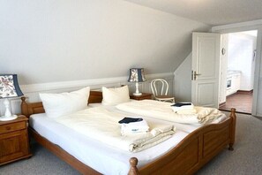 A neatly made bed with a wooden headboard, a bedside table with a lamp, and a chair in a room with white walls and a door leading to another room. - Lindenhof Holiday Apartment 5 (Wenningstedt-Braderup)