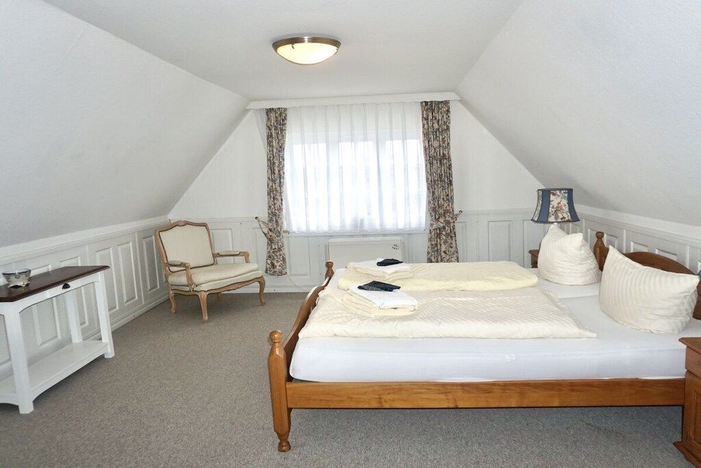 Een slaapkamer met een houten bed, een stoel, een nachtkastje, een lamp en een raam met gordijnen.