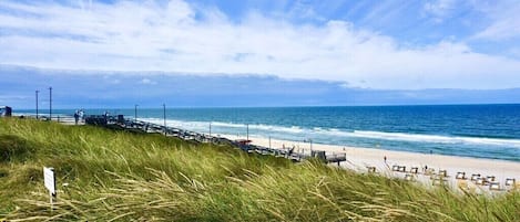 Ein Strand mit einer Holzstege, grasbewachsenen Dünen und dem Meer.