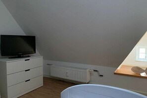 Habitació amb un radiador blanc, un moble blanc amb calaixos, una televisió a sobre i un sostre inclinat.