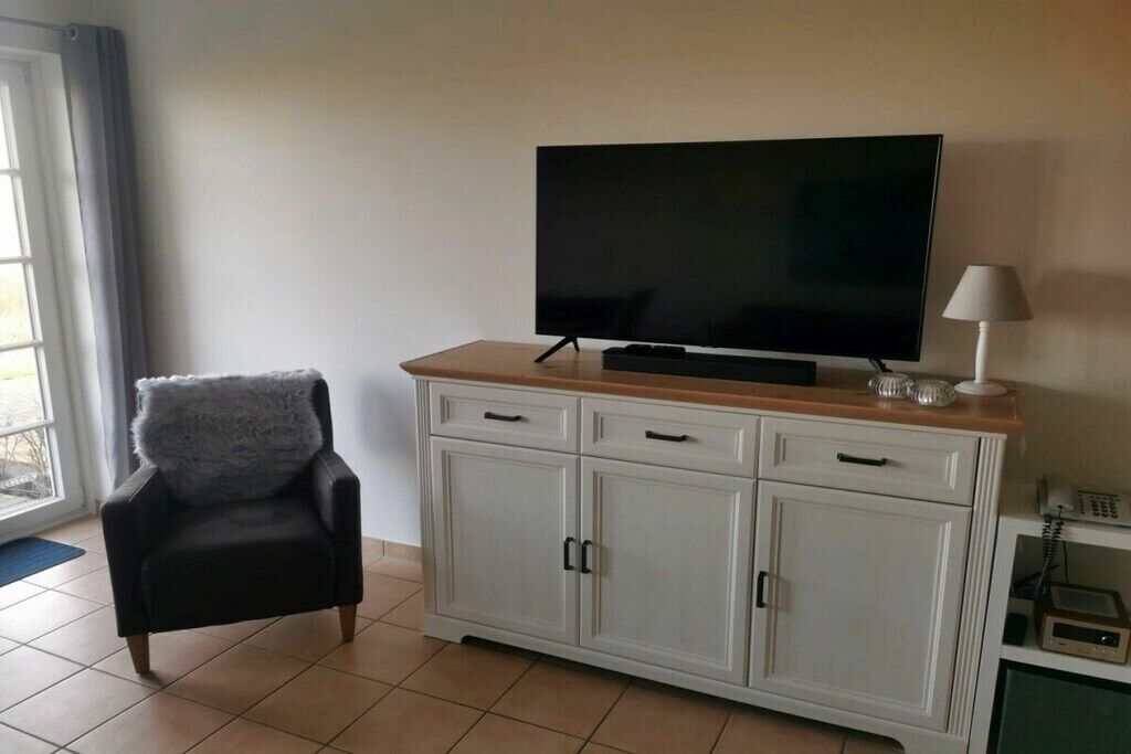 Una sala de estar con un televisor de pantalla plana sobre un mueble de madera, un sillón negro con un cojín mullido y una lámpara blanca sobre una mesita.