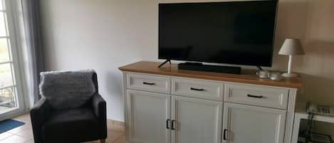 Una sala de estar con un televisor de pantalla plana sobre un mueble de madera, un sillón negro con un cojín mullido y una lámpara blanca sobre una mesita.