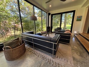 A modern living room with a large window, black leather sofa, and a woven basket. - The Modern | 3 BR Upscale Retreat 3 Blocks to IU (Bloomington)