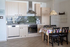 A kitchen with light-colored cabinets, a microwave, a stove, and a coffee maker. There is a dining table with chairs and a hanging light fixture. - Luca Panorama Apt. 107, 8th floor (Cuxhaven)