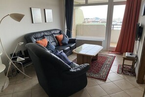A living room with a black leather sofa, a small coffee table, a standing lamp, and a view of the city through a large window. - Luca Panorama Apt. 107, 8th floor (Cuxhaven)