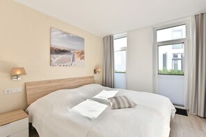 A hotel room with a large bed, a nightstand, a wall-mounted lamp, a window with curtains, and a beach scene painting. - Holiday apartment in Glowe with swimming pool (Glowe)