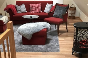 Um sofá vermelho com uma mesa de centro redonda, uma poltrona vermelha, um banquinho cinza com uma manta branca e um abajur preto.
