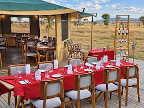 An outdoor dining area with tables set for a meal, a wine rack, and a view of the savanna. - Pori pori Lodges and Camp  (SERENGETI)