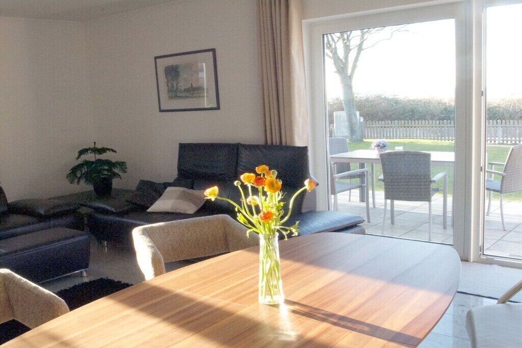 Una sala de estar con una mesa de comedor, un sofá y una planta en maceta. Hay un ventanal corredera que da a un área exterior con una mesa y sillas.