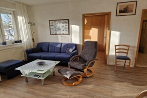 A living room with a blue sofa, a wooden chair, a small table with a lace tablecloth, and a framed picture on the wall. - Travemünde-Priwall - Anchorage (Lübeck Travemünde)