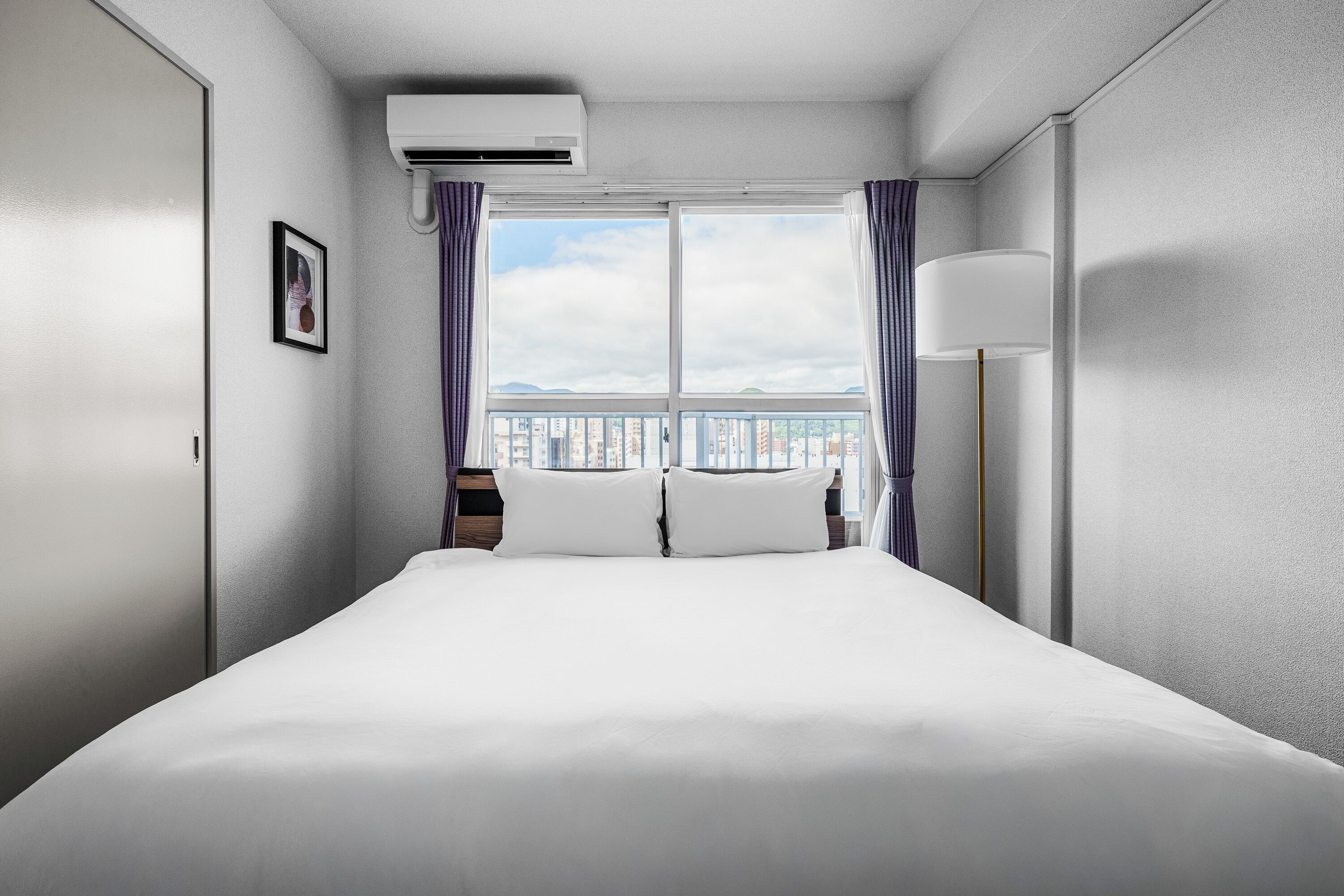 Habitación de hotel con una cama grande, una ventana con cortinas, una mesita de noche y un aire acondicionado de pared.