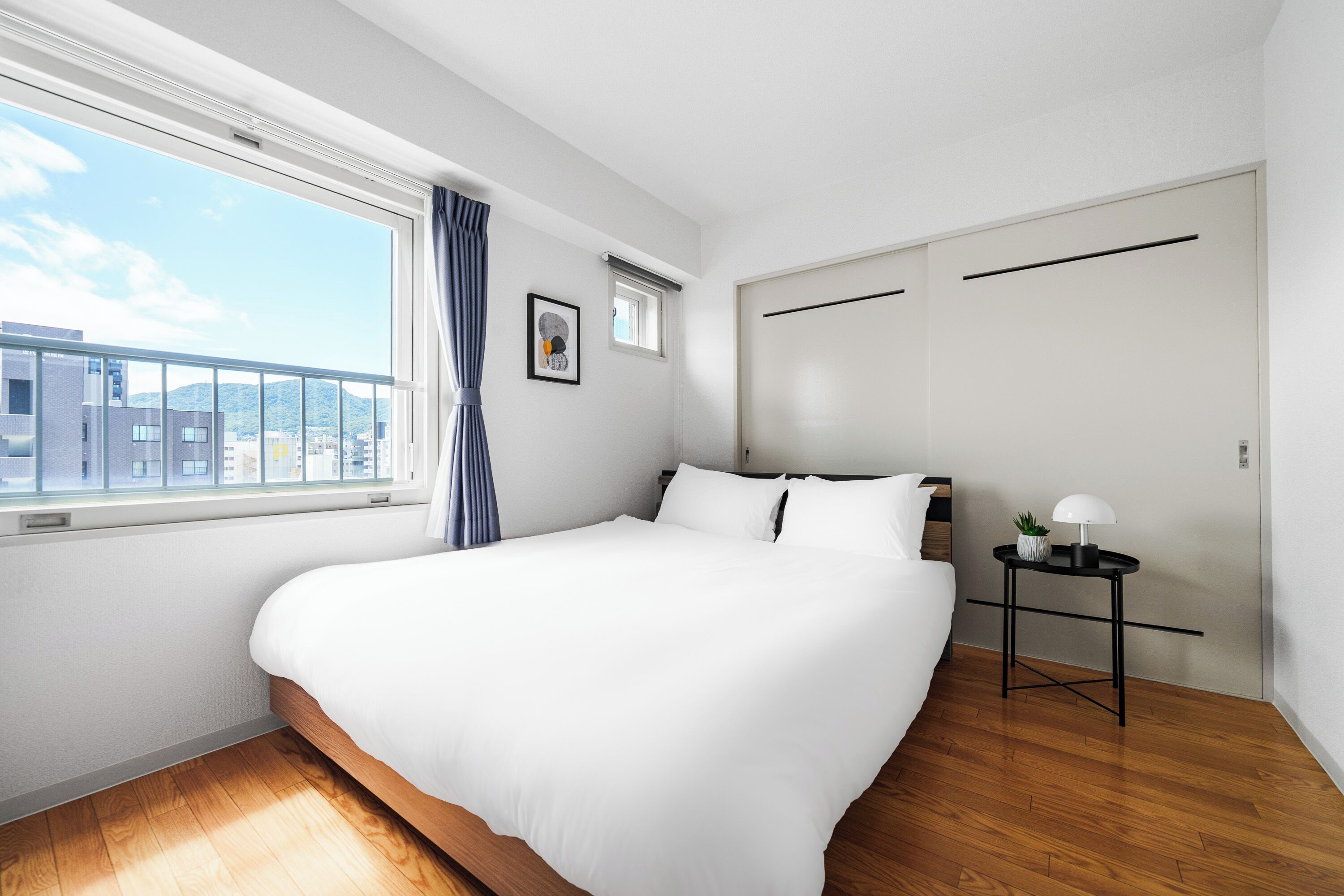 A modern hotel room with a large bed, a window with a view of buildings, a small table with a lamp, and a door.