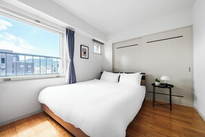 Habitación de hotel moderna con una cama grande, un ventanal con vistas a edificios, una mesita con una lámpara y una puerta.