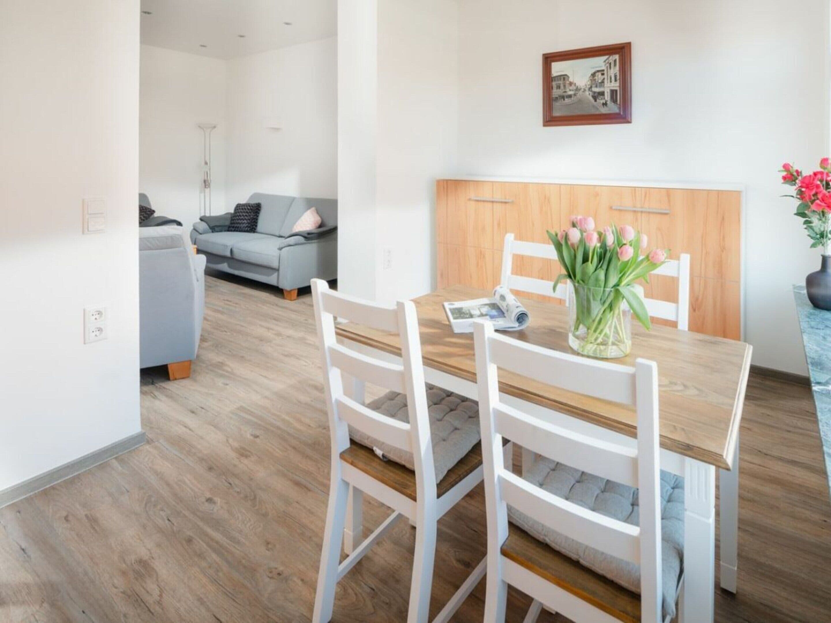 A modern dining area with a wooden table and white chairs, a vase with flowers, and a framed picture on the wall.