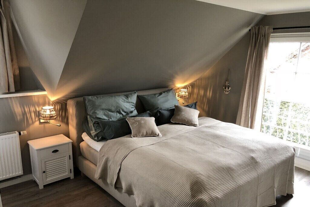 A bedroom with a sloped ceiling, a bed with a beige blanket, two bedside lamps, and a window with curtains.