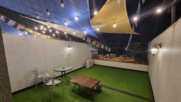 Un patio exterior con césped artificial, una mesa de madera, sillas de metal y luces colgantes.