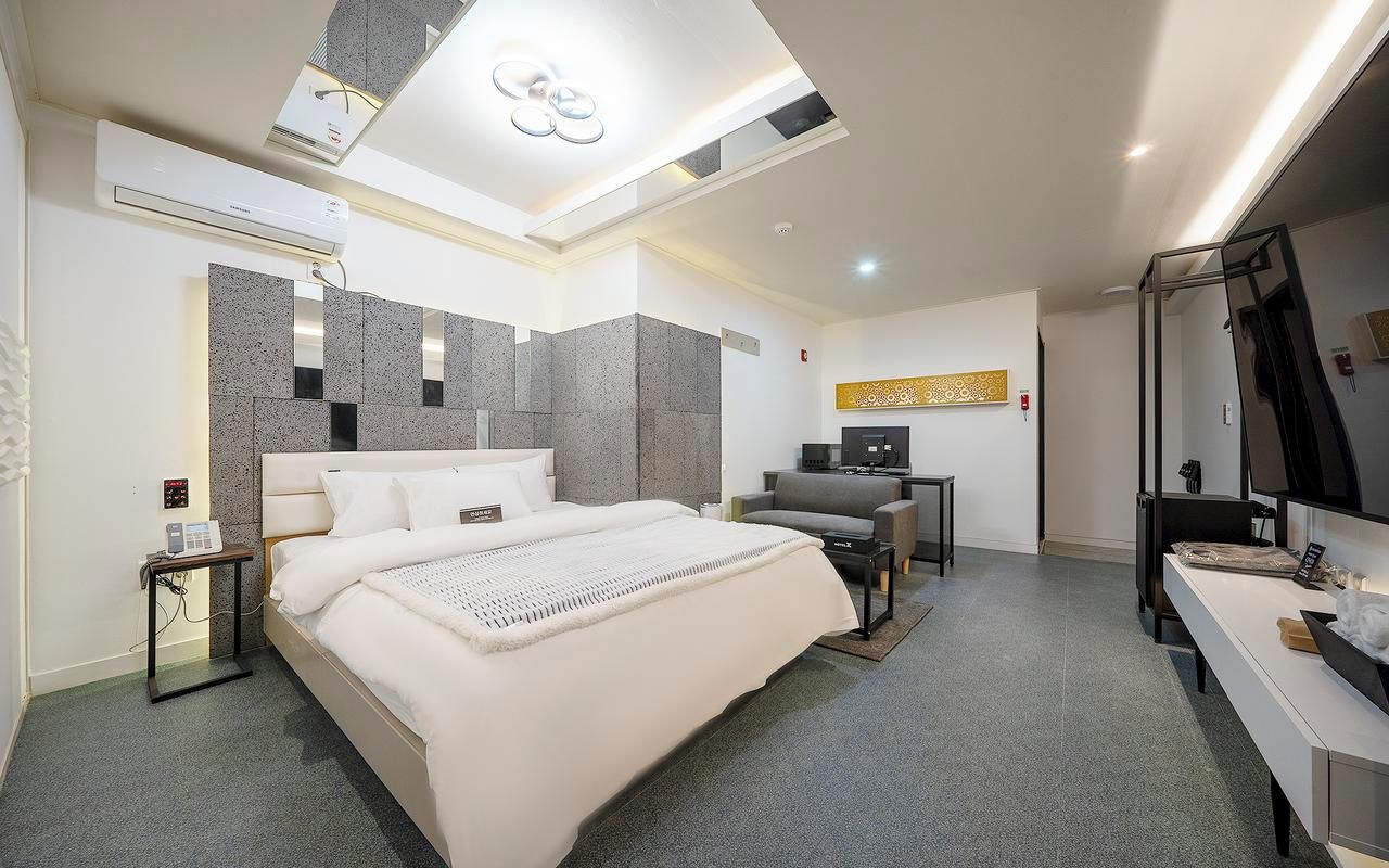 A modern hotel room with a large bed, a desk, a chair, a TV, and a bathroom with a mirror.