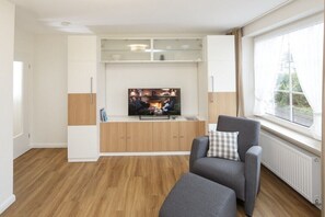 A modern living room with a flat-screen TV, wooden cabinets, a grey sofa, and a large window with a view. - Holiday home Wattenblick 2-room (Cuxhaven)