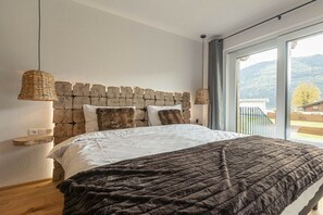 A bedroom with a stone headboard, a large bed with a dark blanket, a wooden bedside table, and a view of the outdoors through a large window. - Superior Holiday Home # 3B with Sauna (Uttendorf)