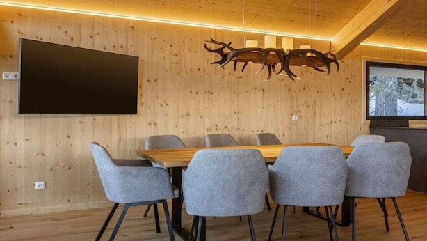 Un comedor moderno con techo de madera, una decoración de cabeza de venado montada, un televisor de pantalla plana y sillas tapizadas de gris alrededor de una mesa de madera.