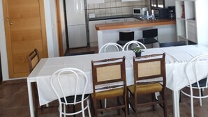 Ruokailutila, jossa on pöytä ja tuolit, keittiö, jossa on kodinkoneet, ja puinen portaikko.