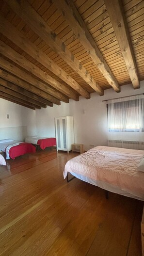 Una habitación con techo de madera, una cama grande, dos camas más pequeñas, un armario y una ventana con cortinas.