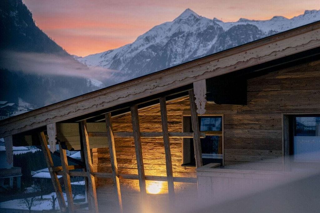 A wooden cabin with a sloped roof, large windows, and a view of snow-capped mountains.