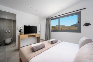 A modern bedroom with a bed, a flat-screen TV, a wooden desk, and a window with a view of hills. - Villa located in Kolympia (Rhodes)