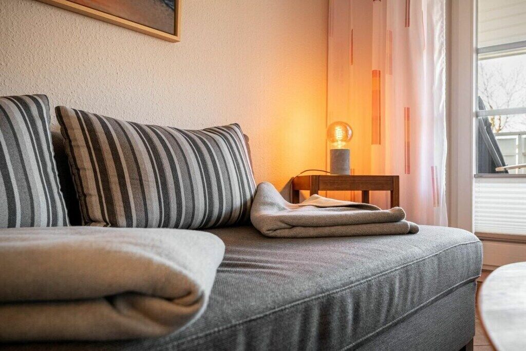 Un sofá con almohadas a rayas y una manta doblada, una lámpara de mesa y una ventana con cortinas.