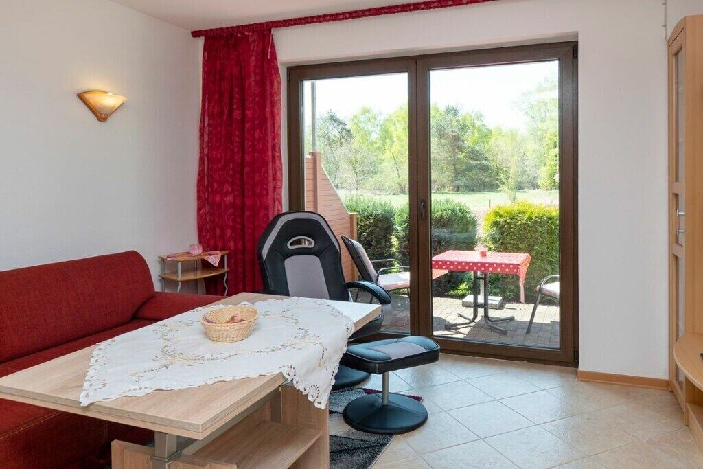 Une pièce avec un canapé rouge, une table recouverte d’une nappe en dentelle, une chaise, une petite table avec une nappe rouge et une porte-fenêtre coulissante donnant sur un espace extérieur équipé d’une table et de chaises.