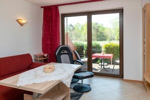 Una stanza con un divano rosso, un tavolo con tovaglia all'inglese, una sedia, un tavolino con tovaglia rossa e una porta scorrevole in vetro che dà su un'area esterna con tavolo e sedie.